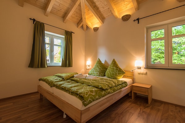Une chambre avec un lit en bois, une literie verte, une table de chevet, une fenêtre avec des rideaux et un plafond en bois.