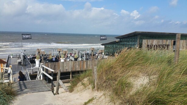 A beachside restaurant with outdoor seating overlooking the ocean, wooden deck access, and a modern building in the background.