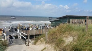 A beachside restaurant with outdoor seating overlooking the ocean, wooden deck access, and a modern building in the background.