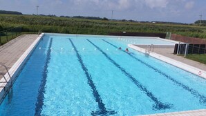 A rectangular swimming pool with multiple lanes, surrounded by a paved deck and a grassy field in the background.