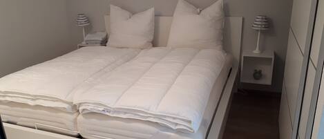 A neatly made bed with white bedding and pillows, a bedside table with a lamp, and a nightstand with a clock.