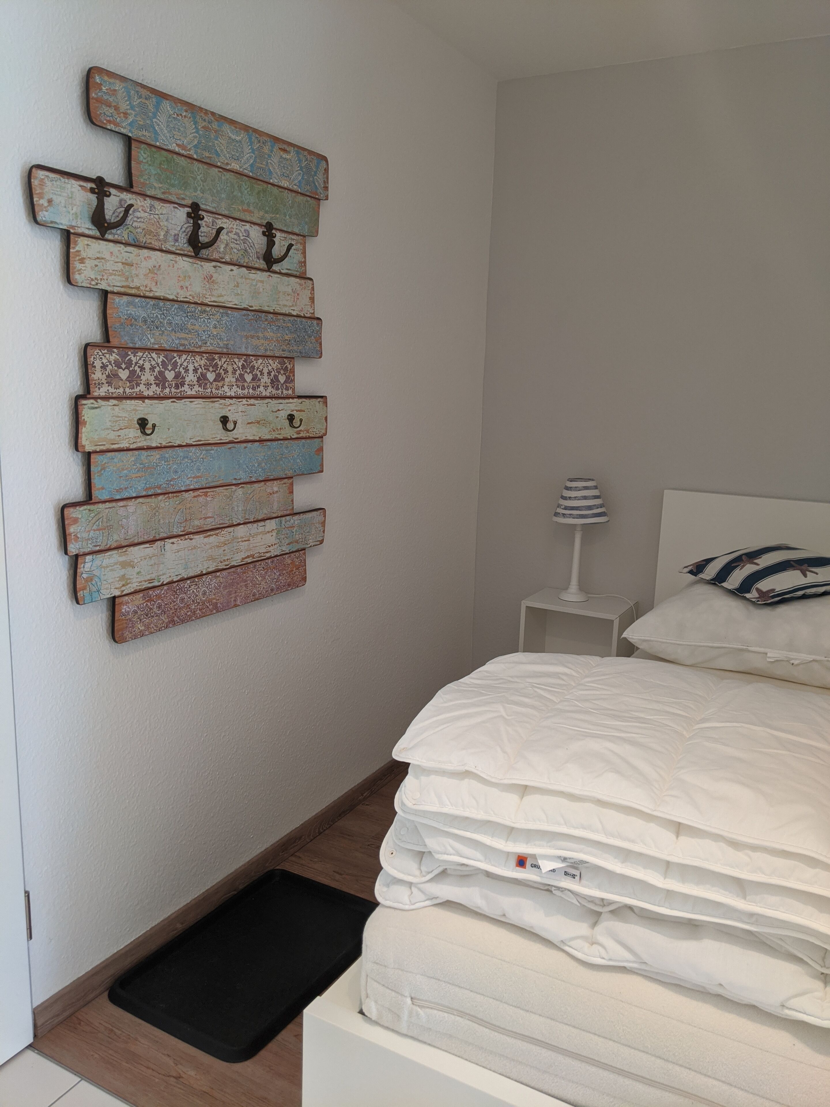 Une chambre à coucher comprenant un lit, une table de chevet, une lampe et un élément décoratif en bois fixé au mur, muni de crochets.