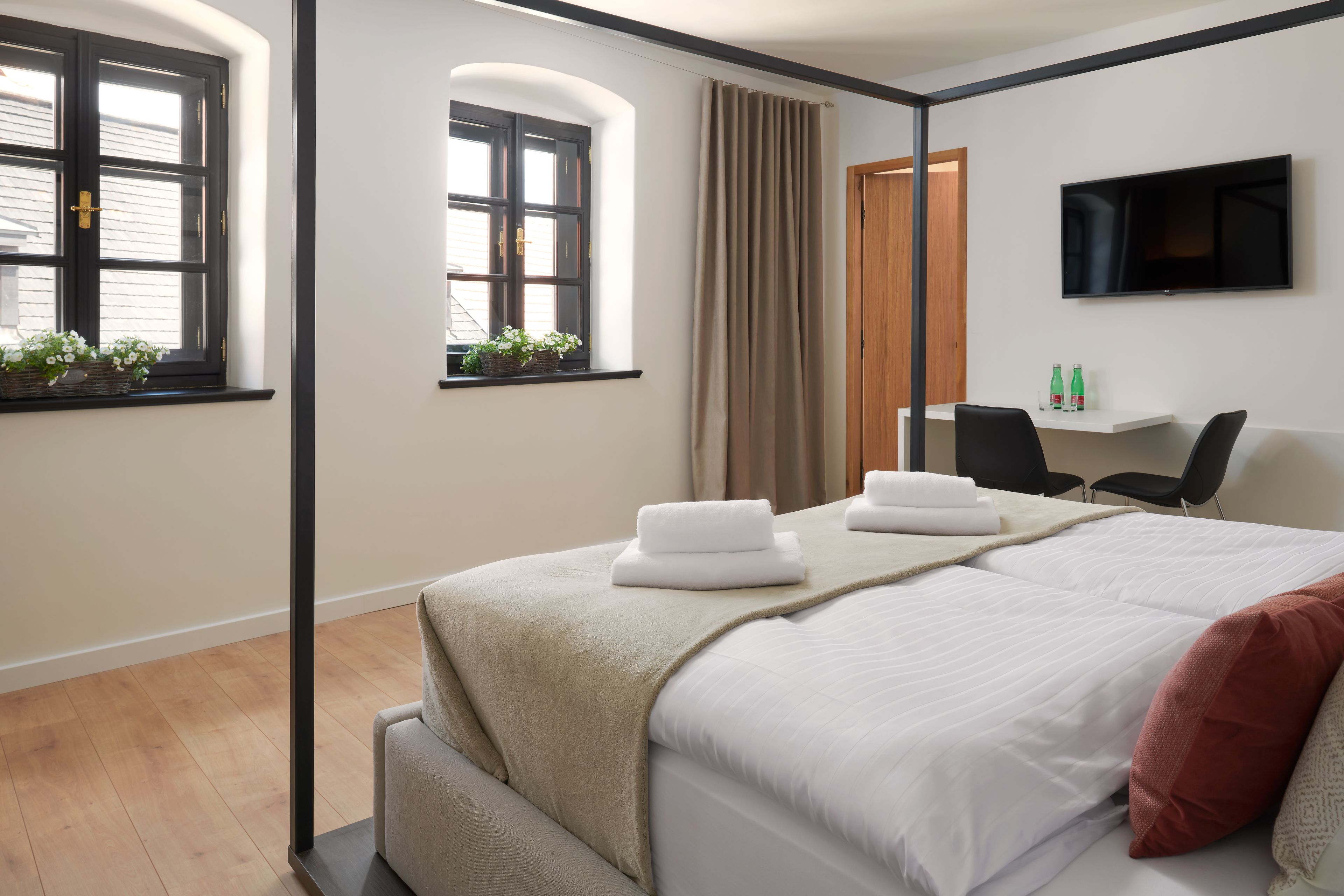 Ein modernes Hotelzimmer mit einem großen Bett, einem Schreibtisch mit Stuhl, einem an der Wand montierten Fernseher und Fenstern mit Vorhängen.