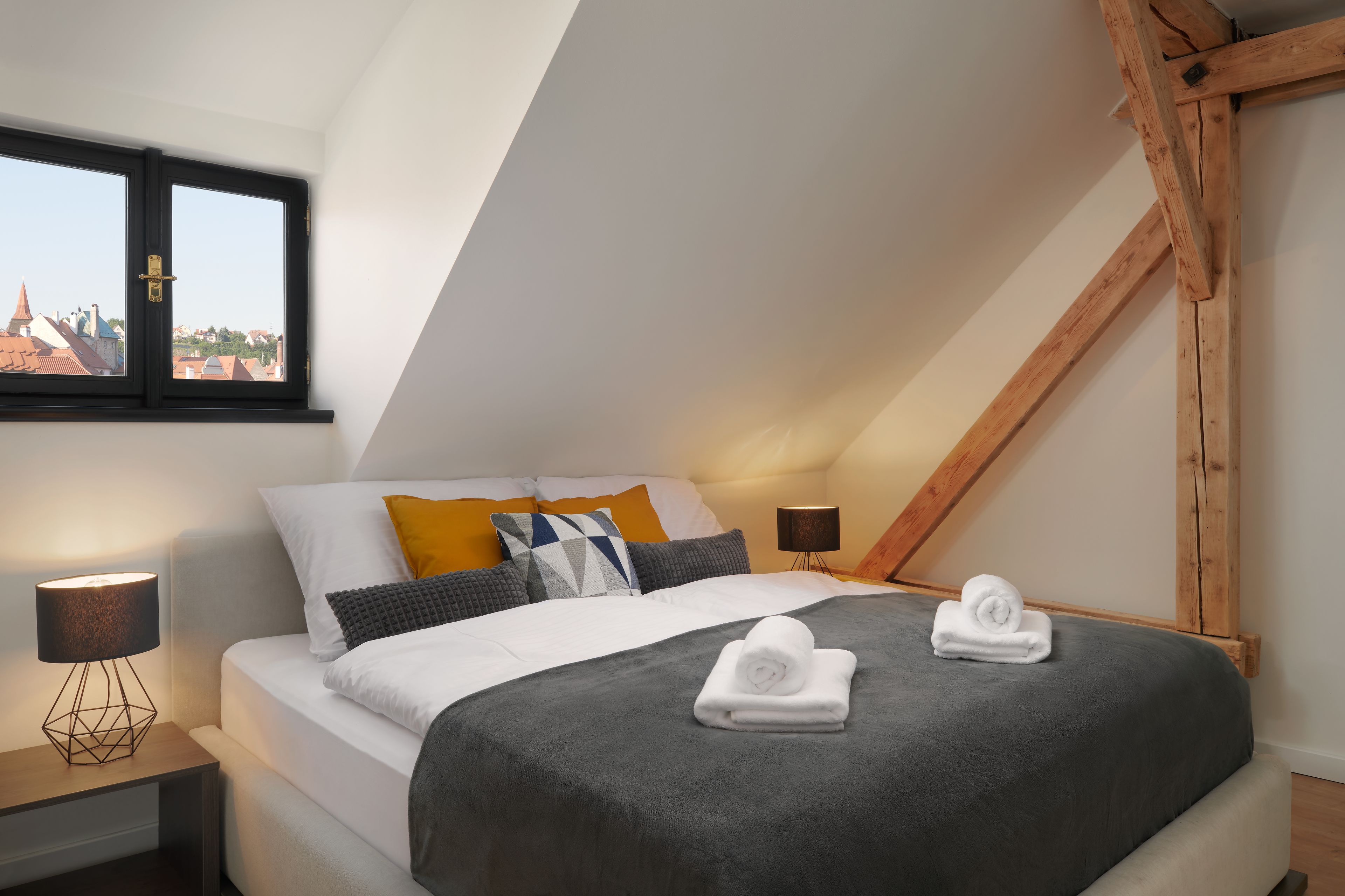 A modern bedroom with a sloped ceiling, a bed with grey bedding, a window with a view of buildings, and a bedside lamp.