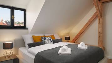 A modern bedroom with a sloped ceiling, a bed with grey bedding, a window with a view of buildings, and a bedside lamp.