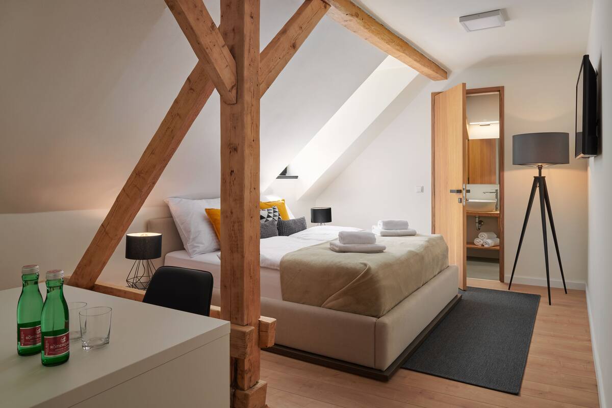 A modern bedroom with a sloped ceiling, a bed with a beige cover, a desk with two green bottles, a lamp, and a tripod-mounted camera.