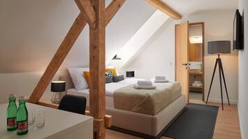 A modern bedroom with a sloped ceiling, a bed with a beige cover, a desk with two green bottles, a lamp, and a tripod-mounted camera.