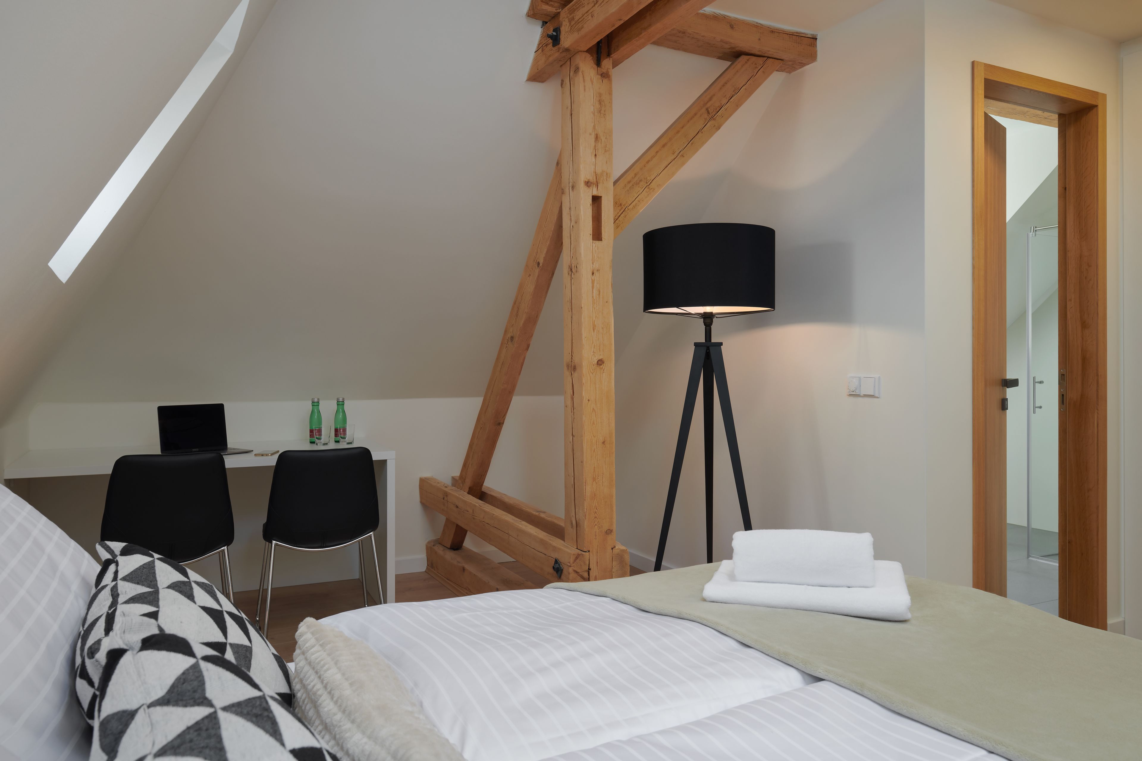 A bedroom with a bed, a desk with two chairs, a lamp, and a wooden ceiling structure.