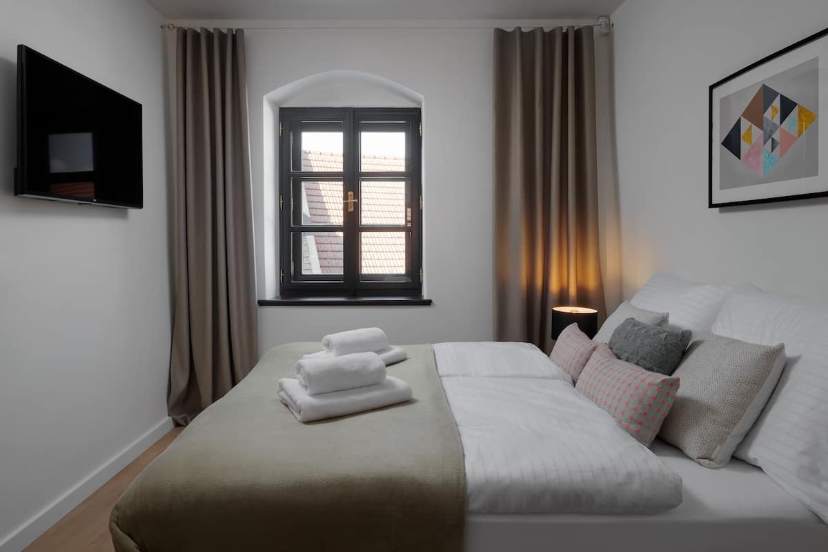 A hotel room with a bed, a TV mounted on the wall, a window with curtains, and a framed picture on the wall.