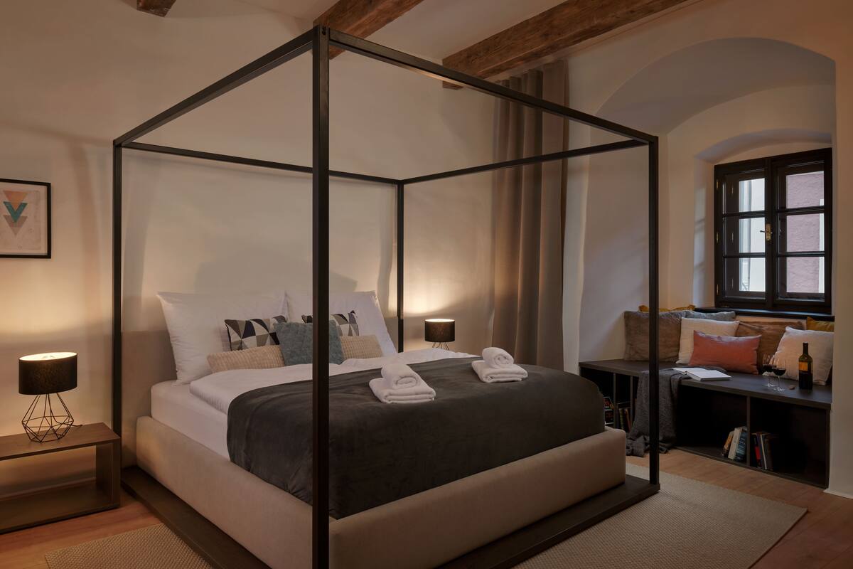 A four-poster bed with a dark bedspread and white linens, a bedside table with a lamp, and a window with curtains.