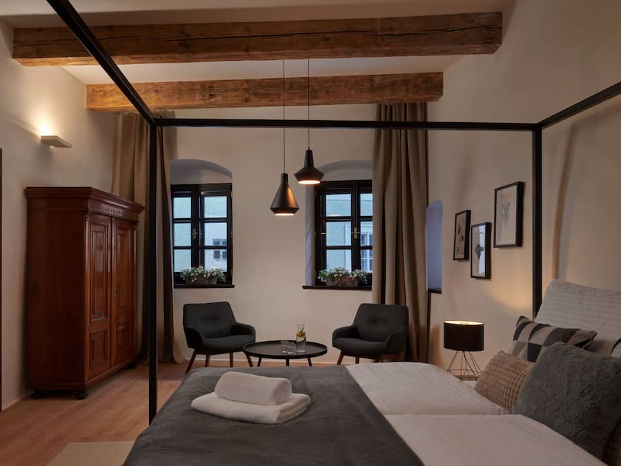A bedroom with a large bed, two chairs, a small table, a wardrobe, and windows with curtains.