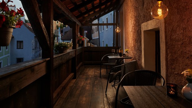 Un balcón acogedor con barandillas de madera, una mesa con sillas, luces colgantes y plantas en macetas.
