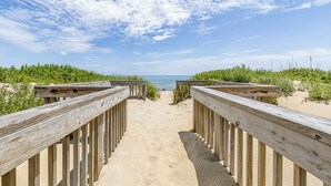 Ein hölzerner Steg führt zu einem Strand mit blauem Himmel und weiter Meerblick.