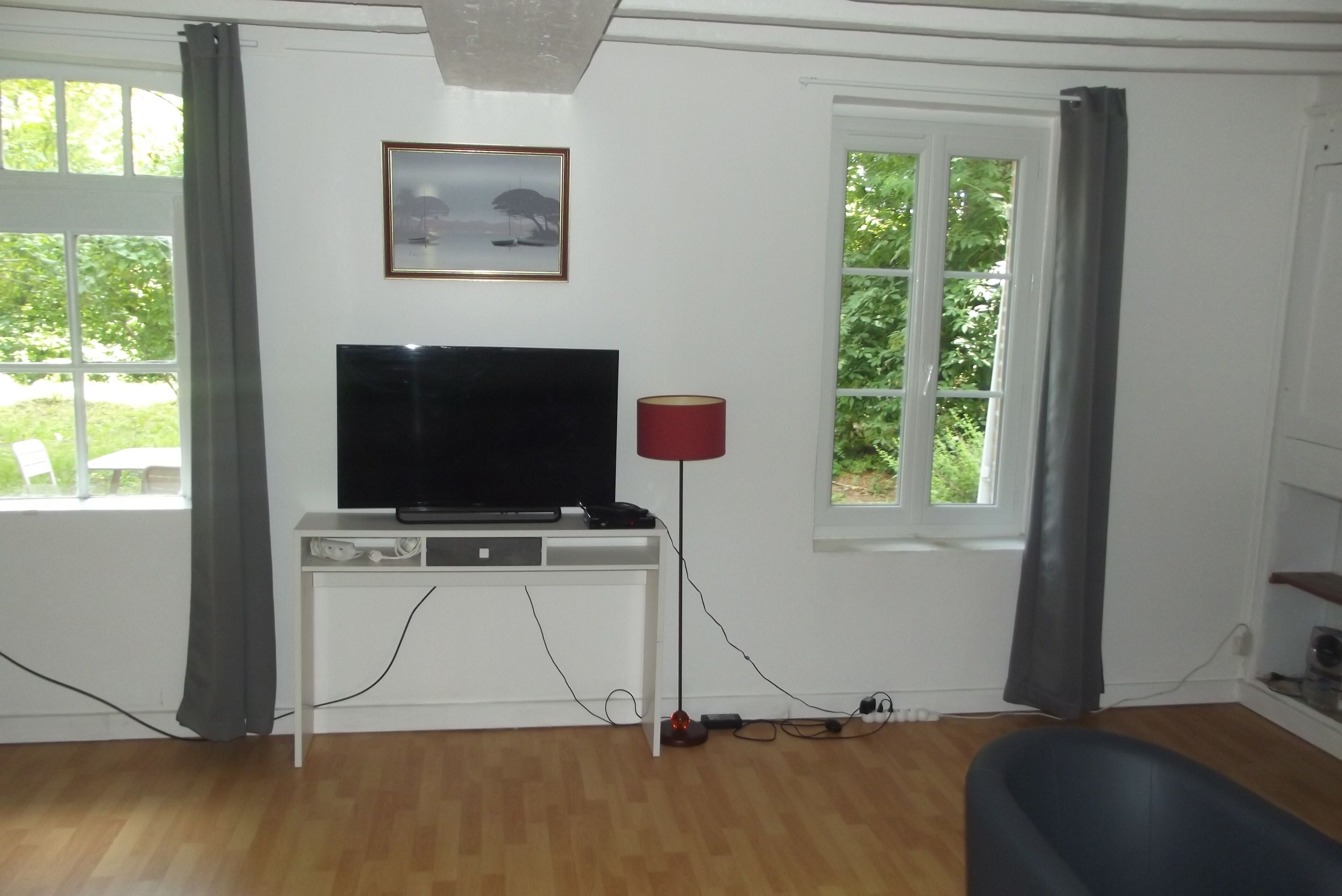 Ein Zimmer mit einem Flachbildfernseher auf einem weißen Standfuß, einer roten Stehlampe und einem Fenster mit Vorhängen.