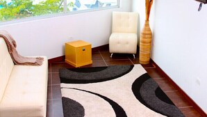 Un salon comprenant un canapé, un petit meuble jaune, un fauteuil blanc, un grand vase et un téléviseur fixé au mur.