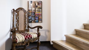 Une chaise en bois avec un coussin à motifs, placée dans un couloir dont le sol est recouvert d’une moquette et qui mène à un escalier.