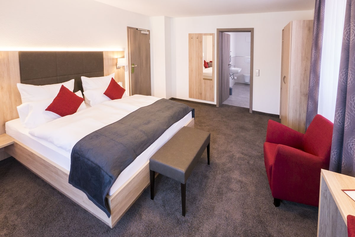 Une chambre d’hôtel moderne avec un grand lit, une banquette, un fauteuil rouge et une armoire en bois.