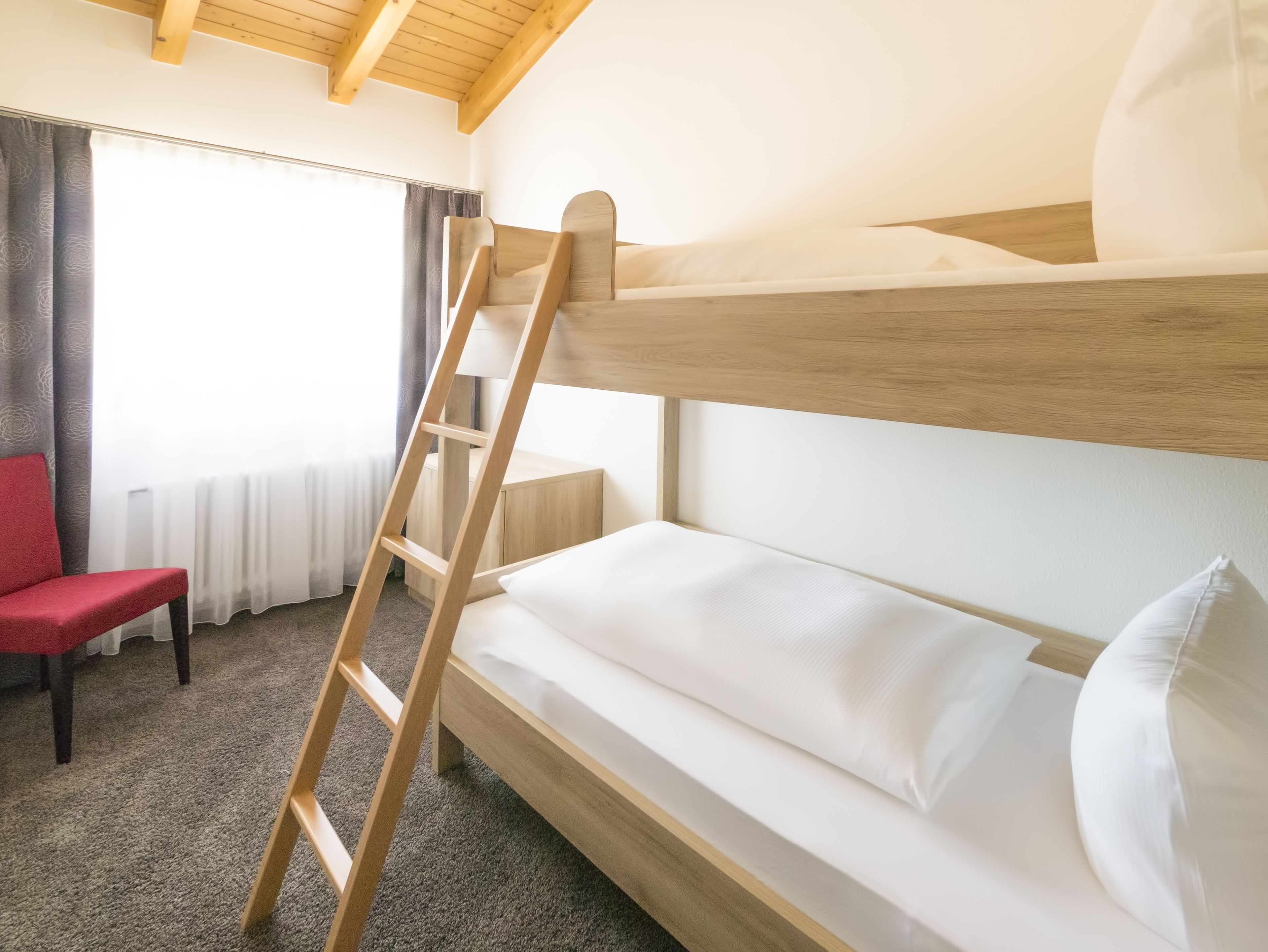 Ein Etagenbett mit einer Holzleiter, ein roter Sessel und ein Fenster mit Vorhängen.