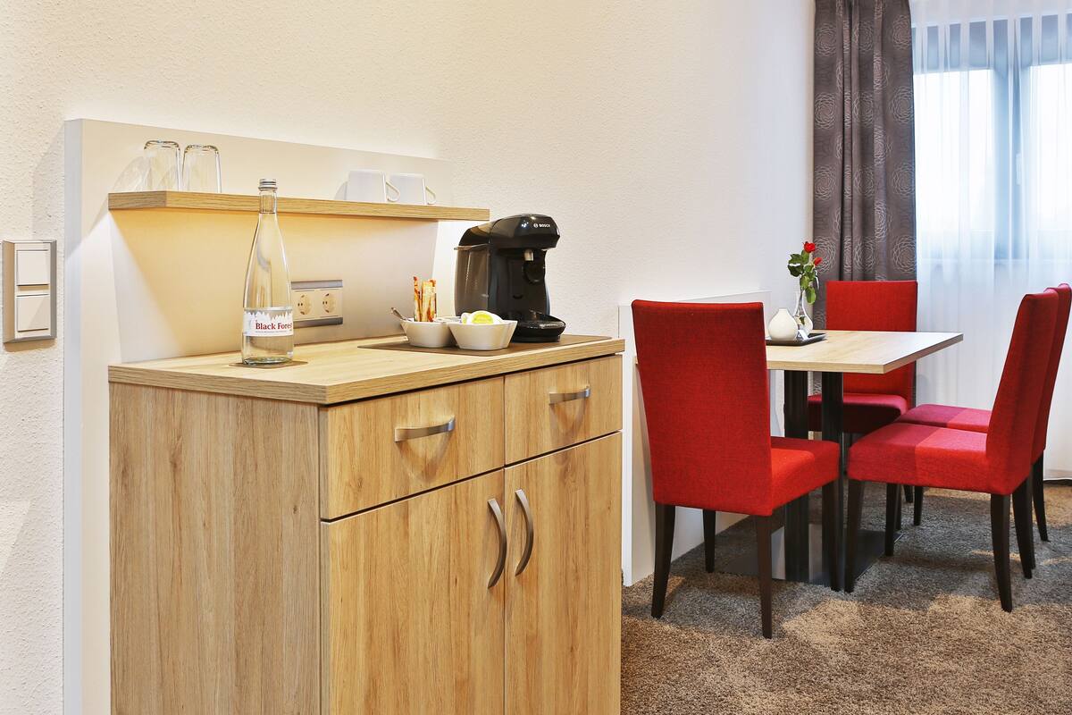 Une cuisine compacte avec un meuble en bois, une machine à café et une petite table entourée de chaises rouges.