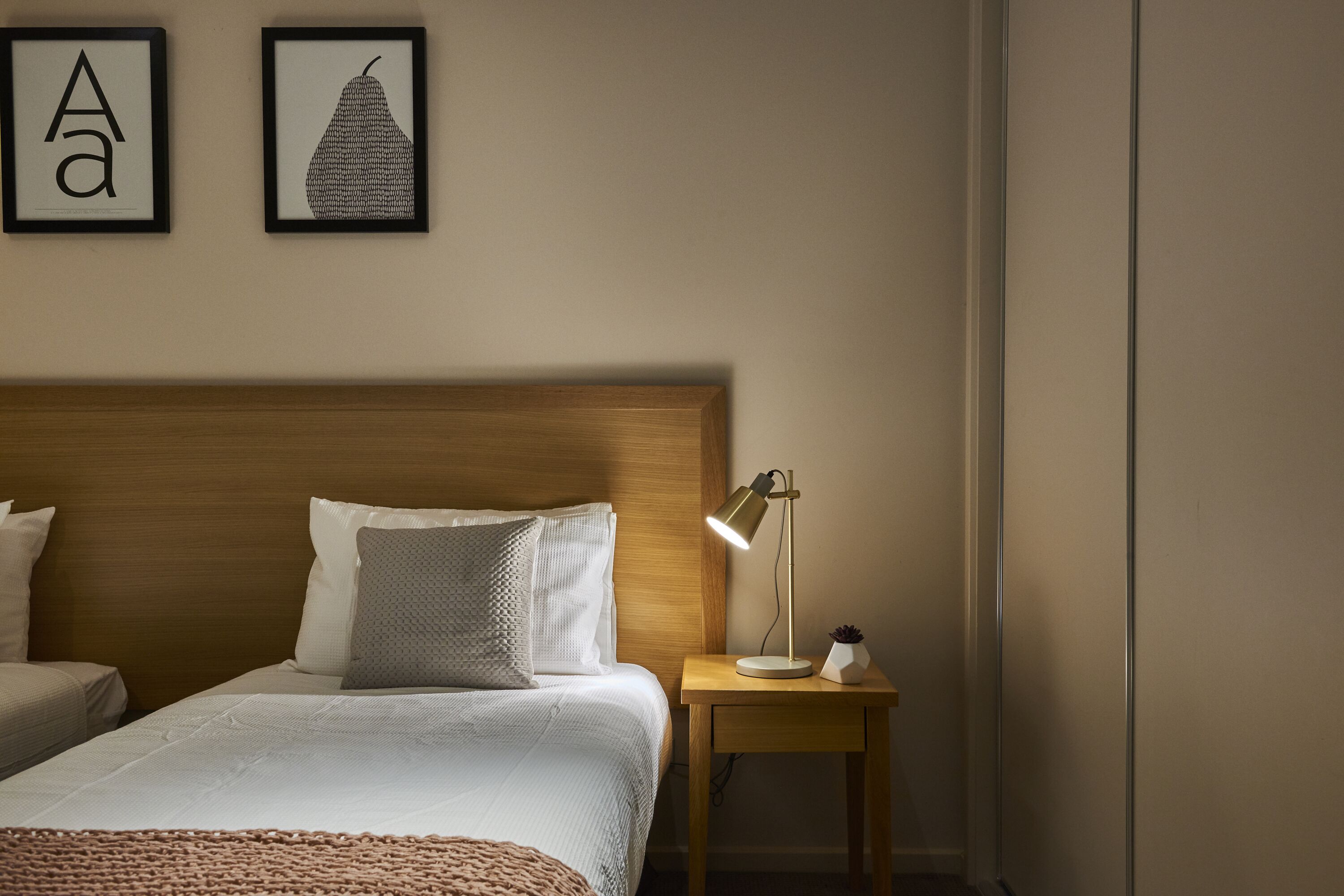 Ein Hotelzimmer mit einem Holzkopfteil, zwei Betten, einem Nachttisch mit Lampe und zwei gerahmten Bildern an der Wand.