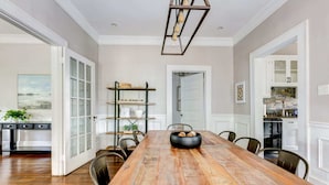 Uma sala de jantar com mesa de madeira, cadeiras, um candelabro e uma cozinha ao fundo.