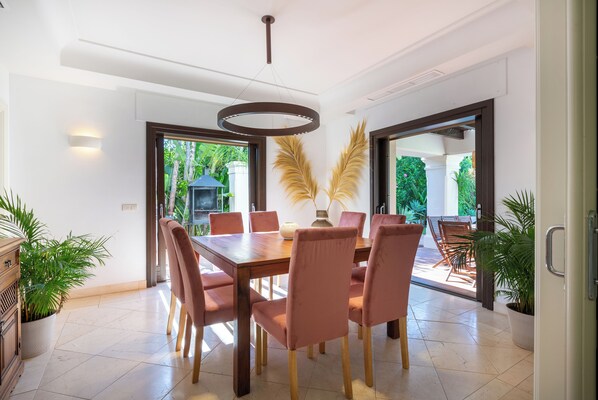 A dining area with a wooden table and six chairs, surrounded by potted plants.
