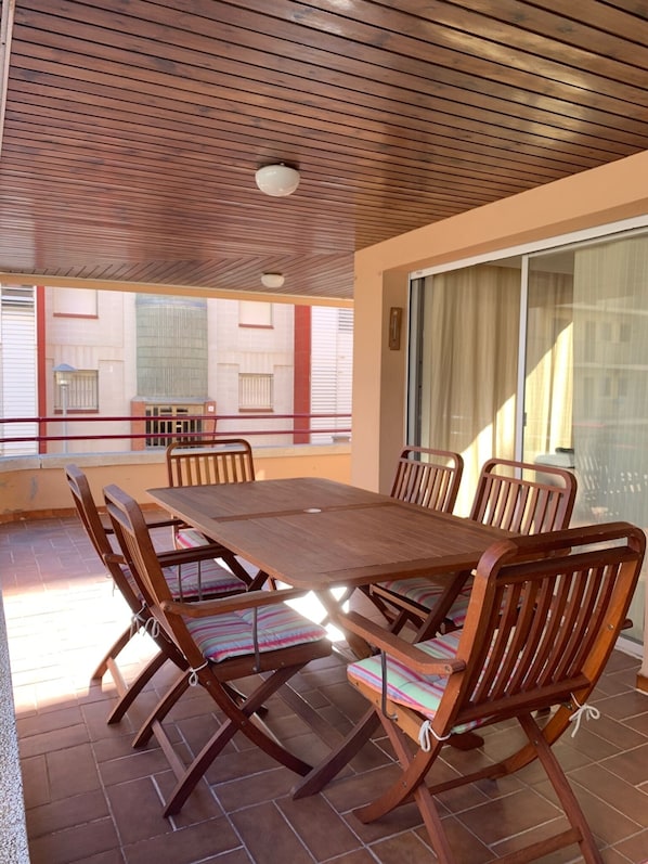 A covered patio with a wooden table and chairs, large windows, and a view of buildings. - Apartment in Torre Valentina, 2 min from the beach (Calonge)