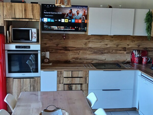 A modern kitchen with a wooden backsplash, a microwave, an oven, a TV mounted above the counter, and a dining area with a table and chairs.