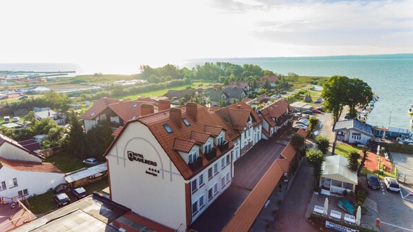 A hotel with a prominent white facade and red-tiled roof, situated by a waterfront with boats docked nearby.