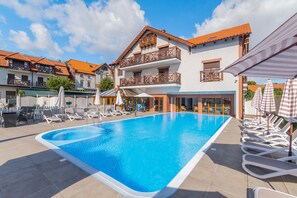 A swimming pool with lounge chairs and umbrellas, surrounded by buildings with balconies and a tiled roof.