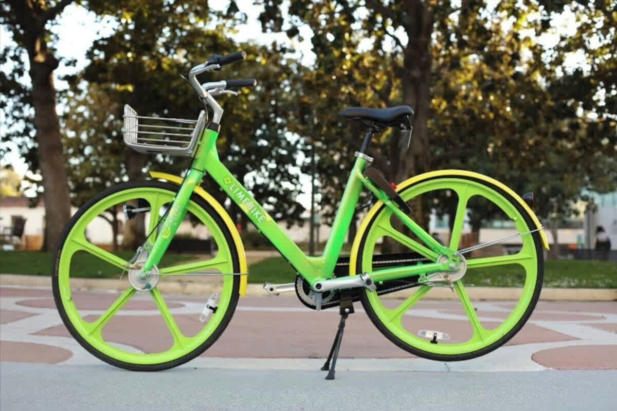 Una bicicleta verde con una cesta y un asiento, estacionada sobre una superficie pavimentada, con árboles al fondo.
