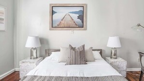 Un dormitorio ordenado con una cama grande, dos mesitas de noche con lámparas, un cuadro en la pared y vistas a un muelle de madera que se adentra en un cuerpo de agua tranquilo.