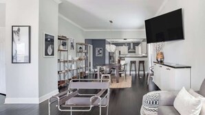 Una sala de estar moderna con un sofá, una mesa de centro y un televisor montado en la pared. Se aprecia una zona de cocina al fondo con una mesa de comedor y sillas.