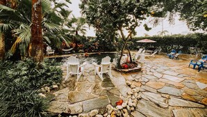 A stone patio with white chairs, a small tree, and a pool area.
