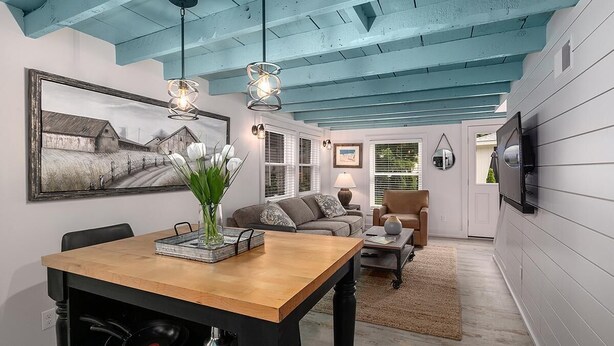 A dining area with a wooden table, chairs, and a vase of flowers. There is a painting on the wall and a television mounted on the wall. - Singapore Stable hot tub walk to Saugatuck! (Saugatuck)