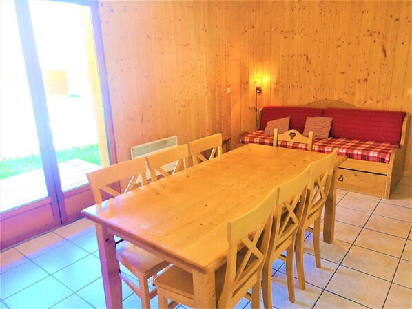 A wooden interior with a dining table, chairs, a sofa with plaid cushions, and a wall-mounted lamp. - Charming Chalet in Les Flocons du Soleil - 3 Rooms, 7 Guests, Pool & Sauna Access (La Joue du Loup)