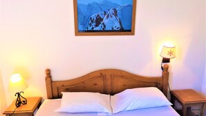 Hotellihuoneessa on puinen sänky, kaksi yöpöytälamppua, kehystetty vuoristomaisema ja pieni pöytä sängyn vieressä.
