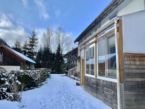 Ein verschneiter Pfad führt zu einem Holzhaus mit großen Fenstern.