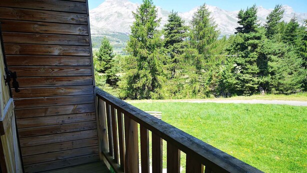 Une terrasse en bois offrant une vue sur un champ verdoyant et des arbres, et s’étendant jusqu’à une chaîne de montagnes.