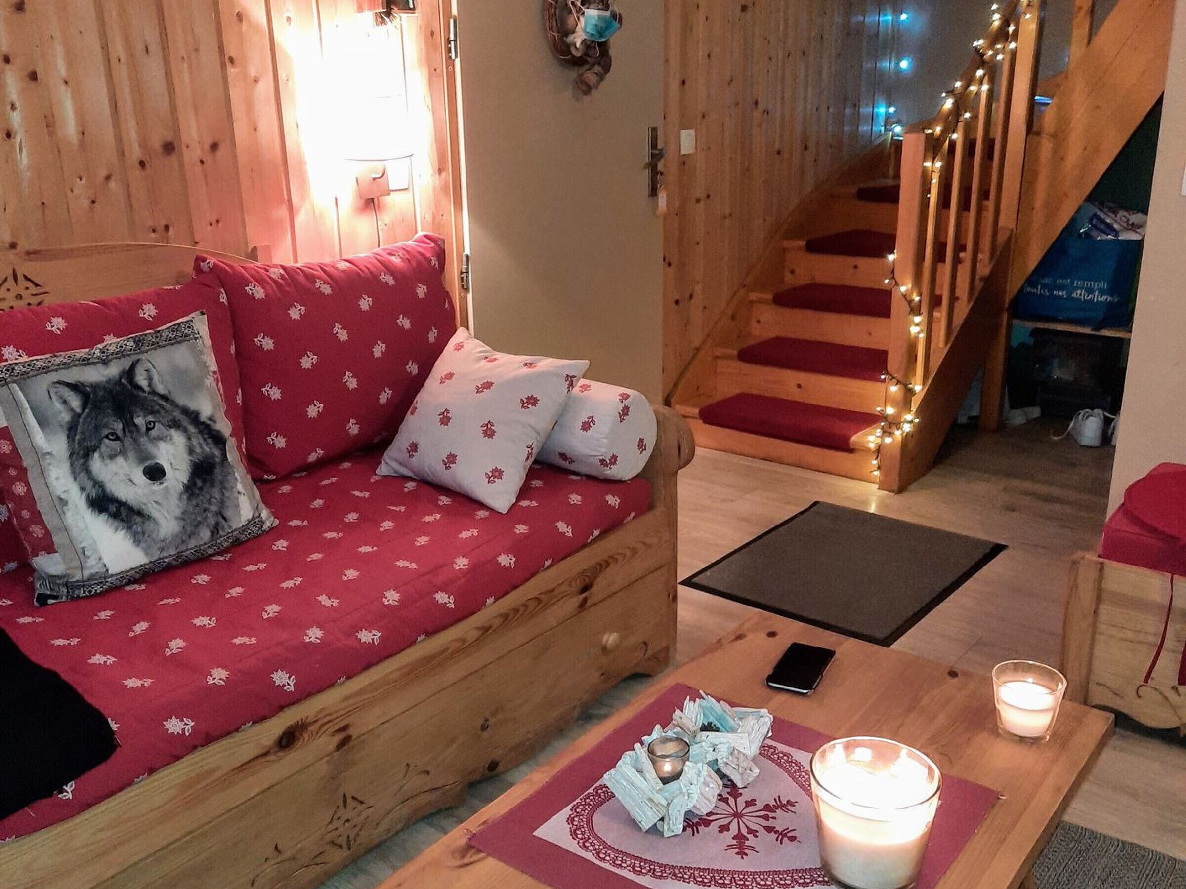 A cozy wooden interior with a red patterned sofa, a wooden staircase with red steps, and a candle on a table.