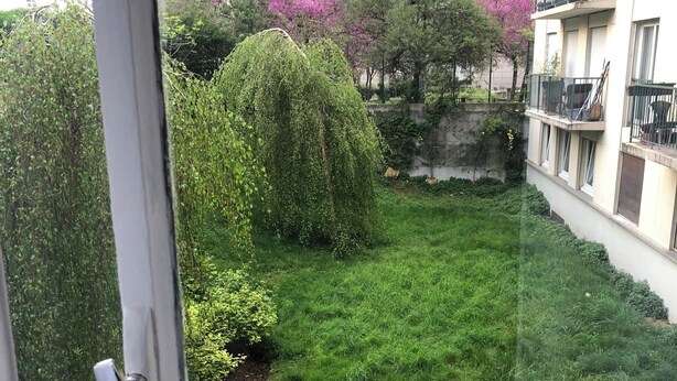 Uitzicht vanaf een balkon over een verzorgde tuin met diverse bomen en struiken, een klein vijvertje en een stenen muur.