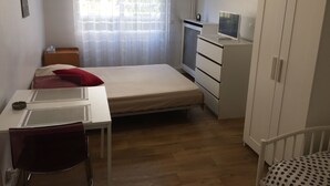 Een kleine, verzorgde kamer met een enkel bed, een bureau met stoel, een garderobe en een raam met gordijnen.