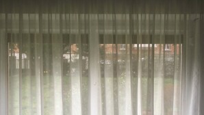 Een raam met dunne witte gordijnen, gedeeltelijk open, toont een uitzicht op een tuin en een gebouw op de achtergrond.