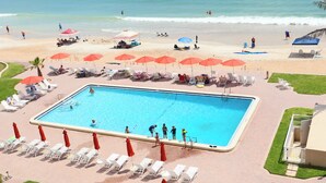 Une piscine avec des chaises longues et des parasols, une plage animée, et une vue dégagée sur l’océan.
