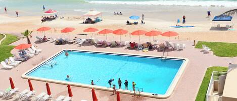 A swimming pool with sun loungers and umbrellas, a beach with people, and a clear view of the ocean.