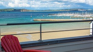 Un balcon avec une chaise rouge, donnant sur une plage où l’on aperçoit des bateaux et un port de plaisance.