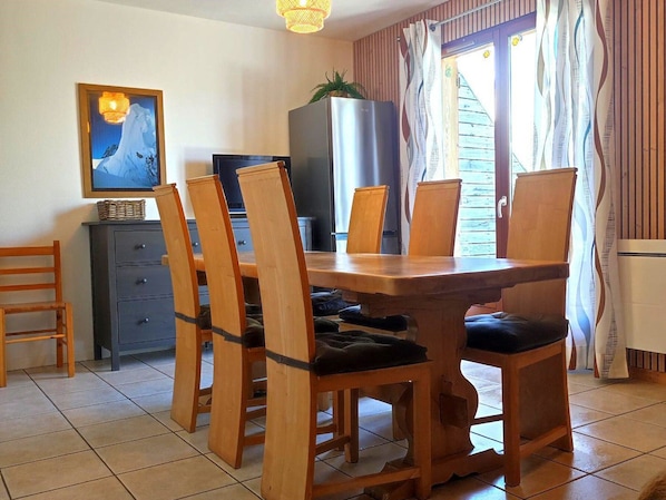 A dining area with a wooden table and chairs, a television, a refrigerator, and a window with curtains. - Cozy Chalet, Sleeps 7, Pool & Sauna Access, Near Shops & Restaurants (Le Dévoluy-Agnières-en-Dévoluy)