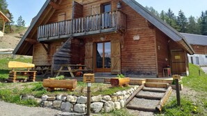 A wooden cabin with a balcony, a staircase, and a stone foundation. - Cozy Chalet, Sleeps 7, Pool & Sauna Access, Near Shops & Restaurants (Le Dévoluy-Agnières-en-Dévoluy)