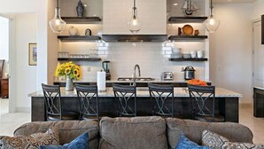 A modern kitchen with a central island, bar stools, pendant lighting, and a dining area with a sofa and patterned cushions.