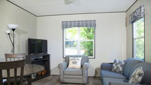 Uma sala de estar com um sofá cinza, uma poltrona cinza, uma TV sobre um móvel, uma mesa de jantar com cadeiras e uma janela com cortina estampada.